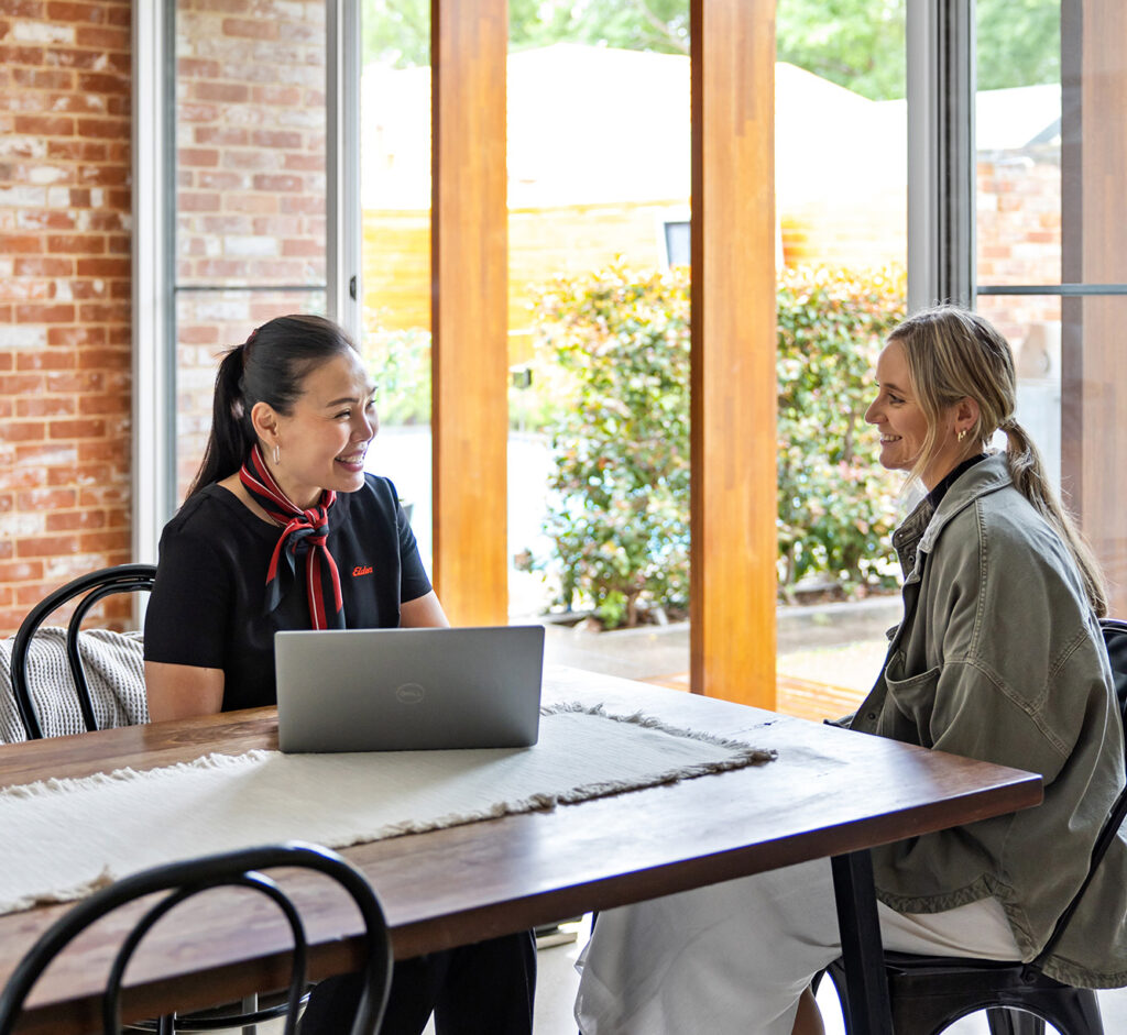 2 women sitting at a dining table talking. 1 has a laptop in front of her